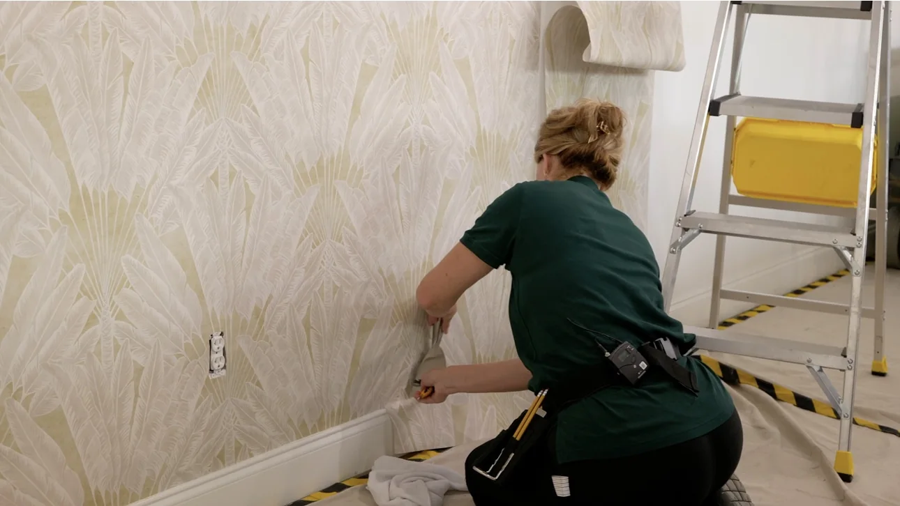 Installer smoothing a pale leaf-pattern wallpaper along the lower edge of a bright room
